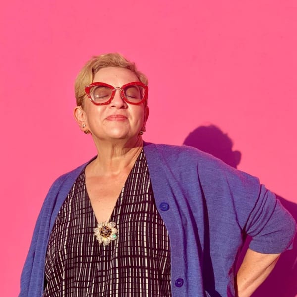 A woman with red glasses and a blue cardigan stands against a bright pink background.