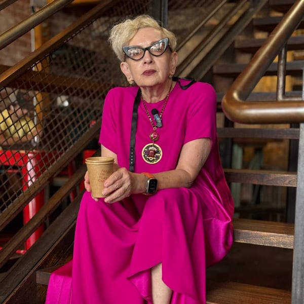 A woman in a vibrant pink dress and cat-eye glasses sits on wooden stairs, holding a Paper or Plastik Cafe cup.
