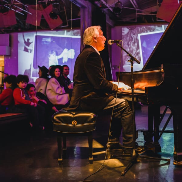 A man with white hair sings into a microphone while playing a grand piano for an audience.