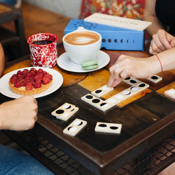Two people playing dominoes at a cafe table with coffee, a raspberry tart, and a macaron.