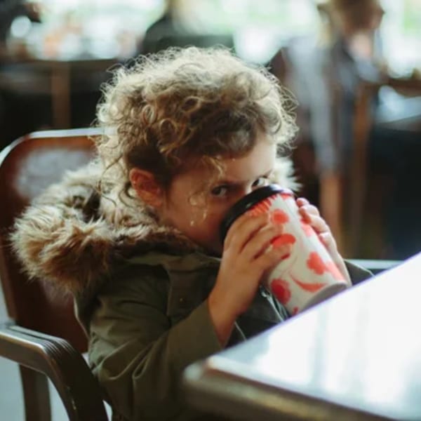 A young child with curly hair sips from a patterned cup, wearing a fur-lined jacket at Paper or Plastik Cafe.