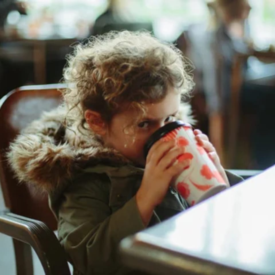 A young child with curly hair sips from a patterned cup, wearing a fur-lined jacket at Paper or Plastik Cafe.