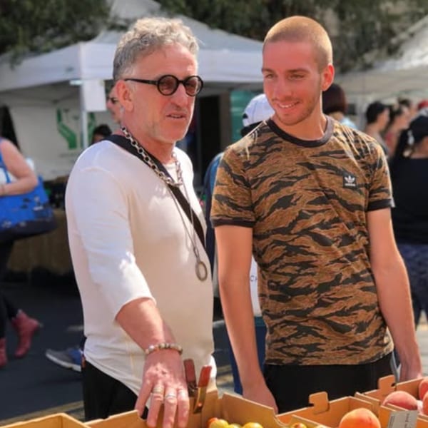 Two men, one older with glasses and one younger, standing by a fruit stand at an outdoor market.