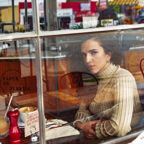 A woman with curly hair sits inside Paper or Plastik Cafe, seen through a window reflection.