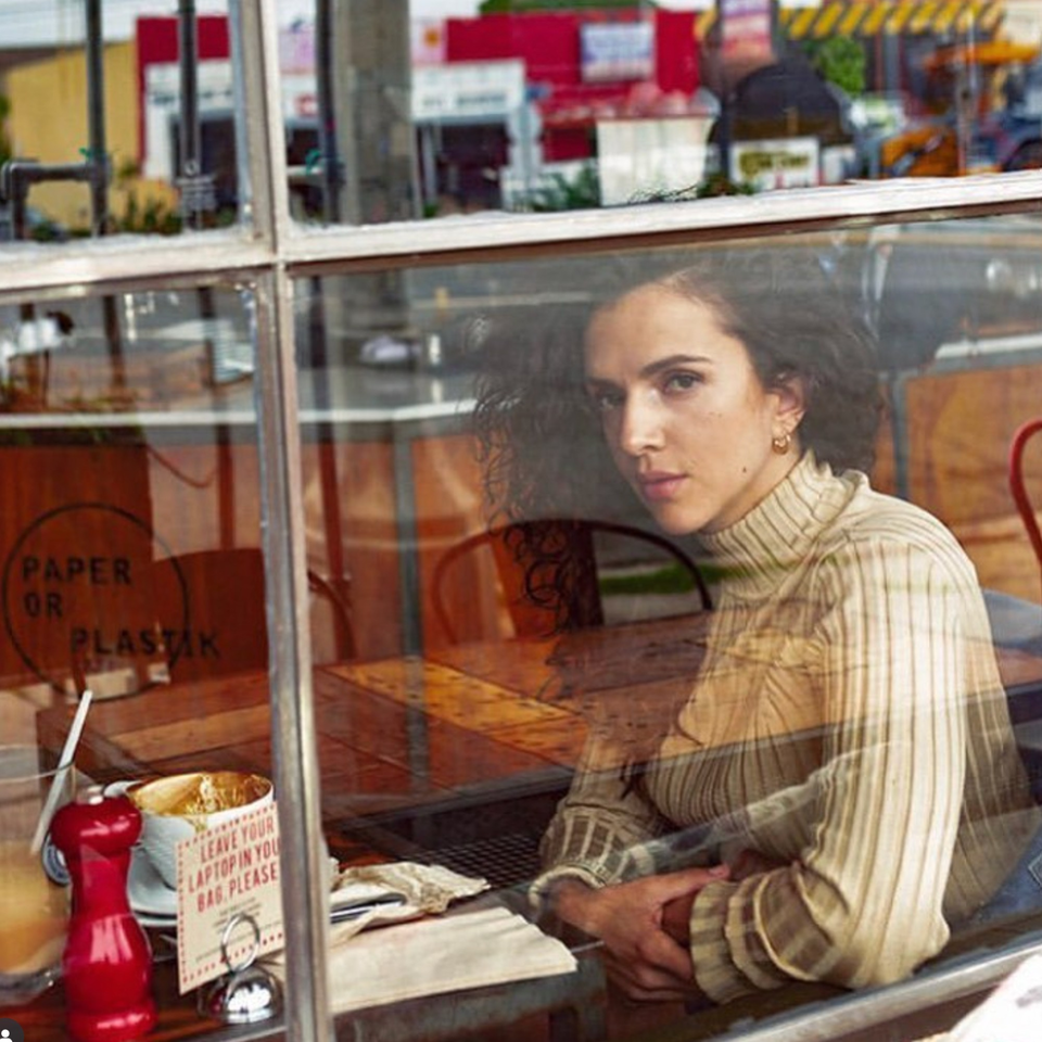 A woman with curly hair sits inside Paper or Plastik Cafe, seen through a window reflection.