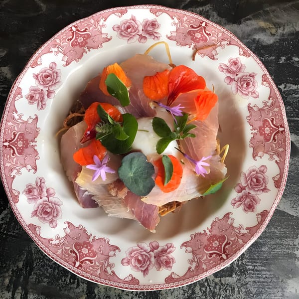 Prosciutto, poached egg, and edible flowers on a decorative white and red plate.