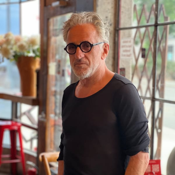 A man with gray hair and round glasses stands in a cafe, looking directly at the camera.
