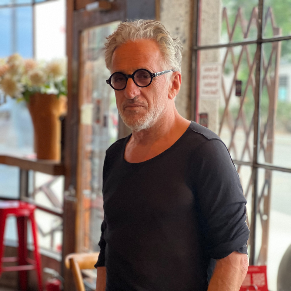A man with gray hair and round glasses stands in a cafe, looking directly at the camera.