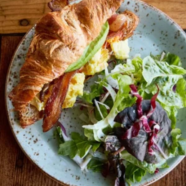 A croissant sandwich with scrambled eggs and bacon, served with a side salad on a blue plate.