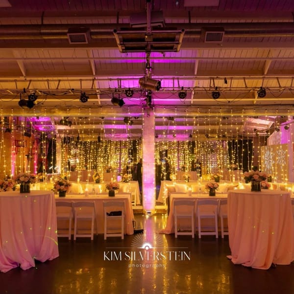 A large event space with tables, chairs, string lights, and pink and yellow uplighting.