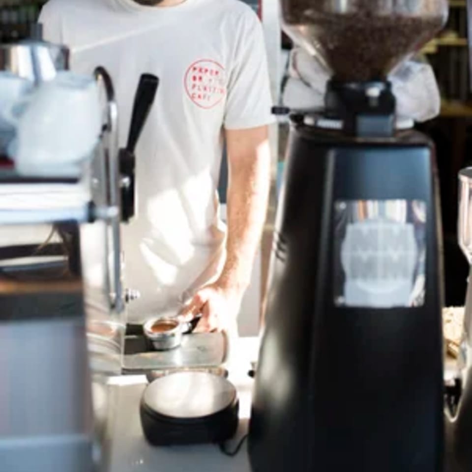 Barista wearing a Paper or Plastik Cafe shirt preparing coffee with an espresso machine and grinder.