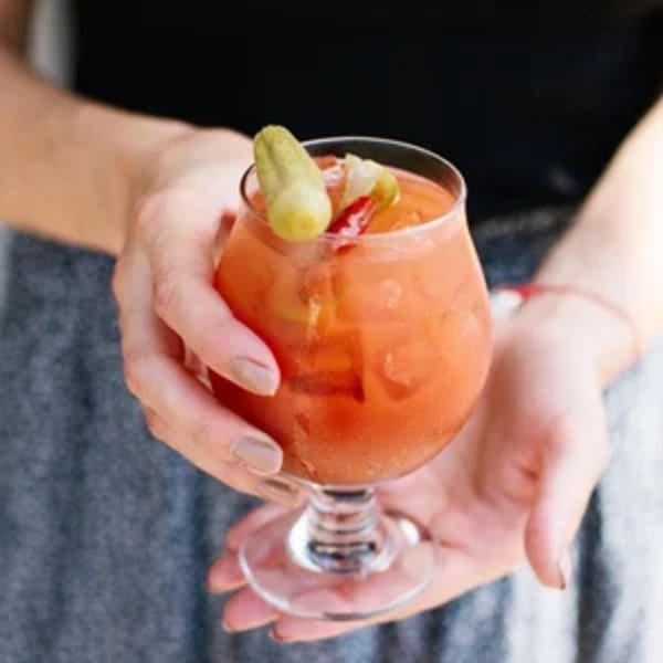 Hands holding a glass of orange drink garnished with a pickle and red pepper.
