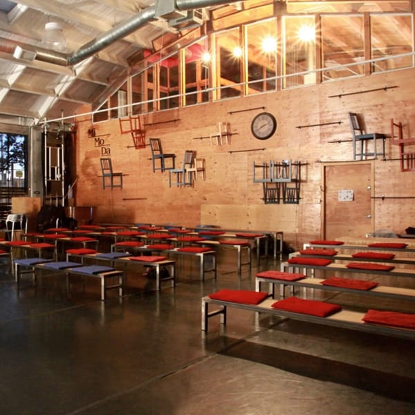 Industrial-style cafe interior with rows of benches, red and blue cushions, and chairs mounted on a wooden wall.