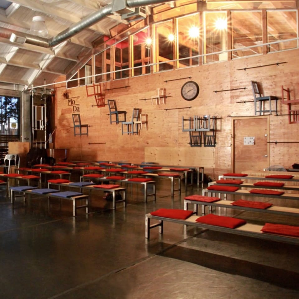 Industrial-style cafe interior with rows of benches, red and blue cushions, and chairs mounted on a wooden wall.