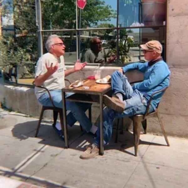 Two men conversing at an outdoor table with drinks and food at Paper or Plastik Cafe.