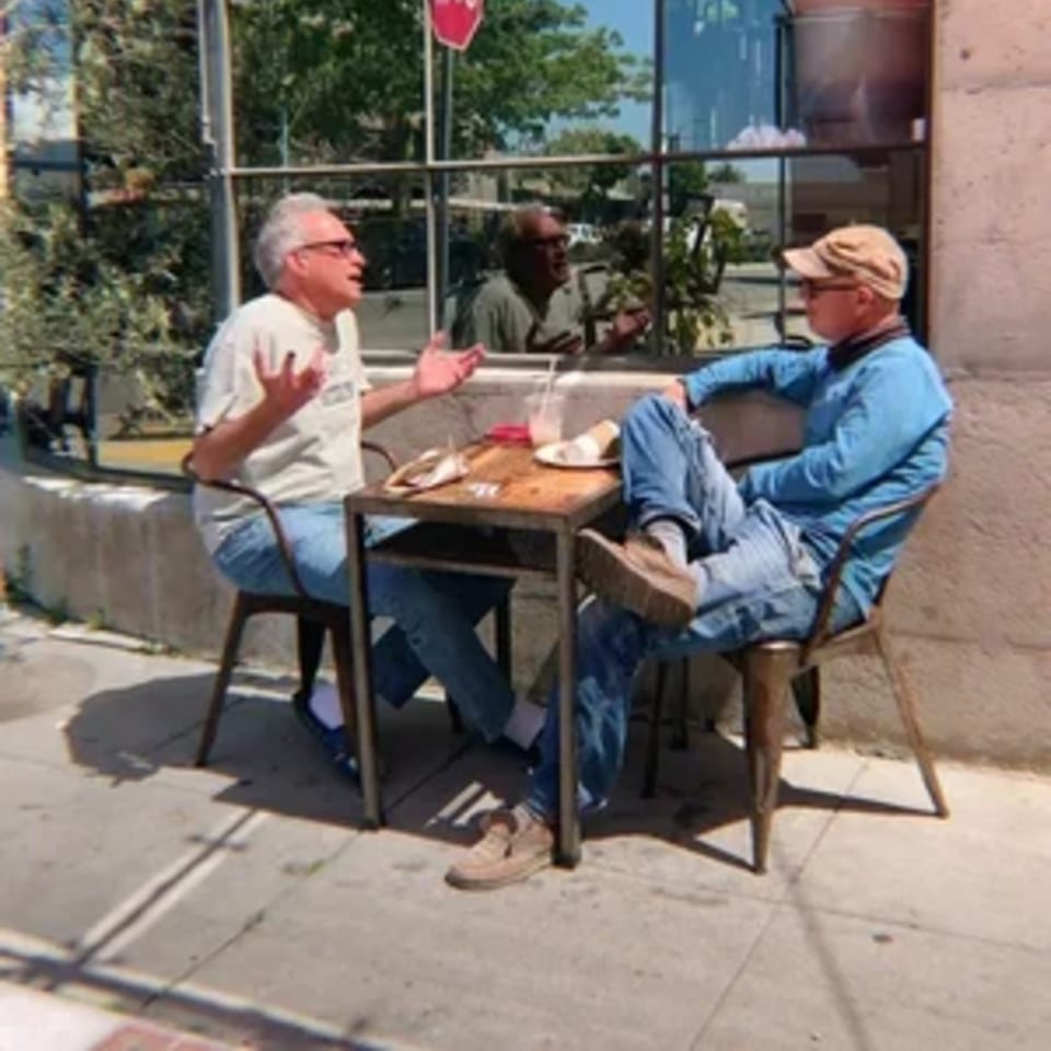 Two men conversing at an outdoor table with drinks and food at Paper or Plastik Cafe.