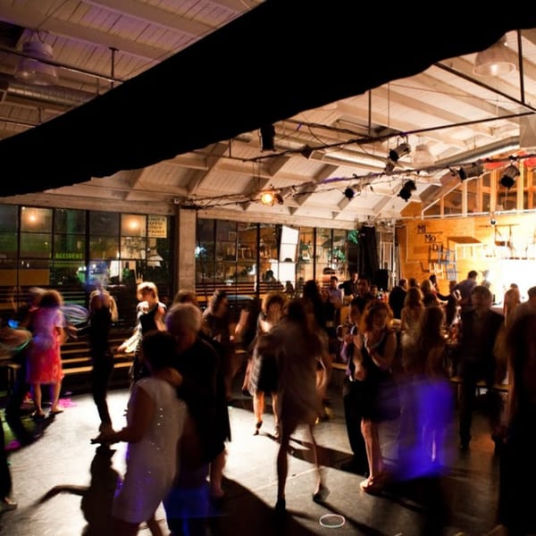 People dancing in a dimly lit, spacious room with large windows and exposed ceiling beams.
