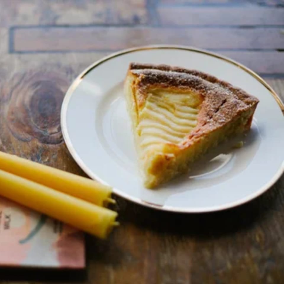 A slice of apple tart with powdered sugar on a white plate with gold rim, next to two yellow candles.