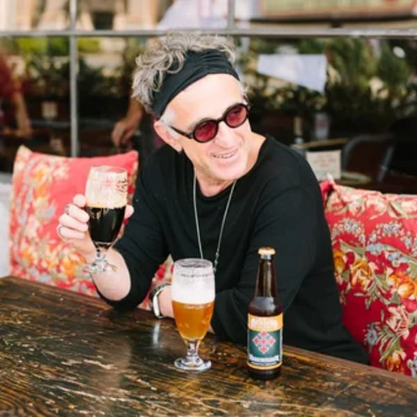 A man in sunglasses and a headband smiles, holding a dark beer at a wooden table.