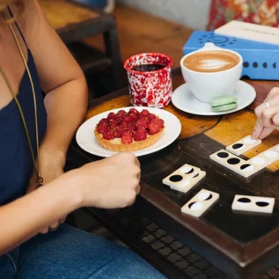 Two people playing a board game at Paper or Plastik Cafe with coffee, a macaron, and a raspberry tart.