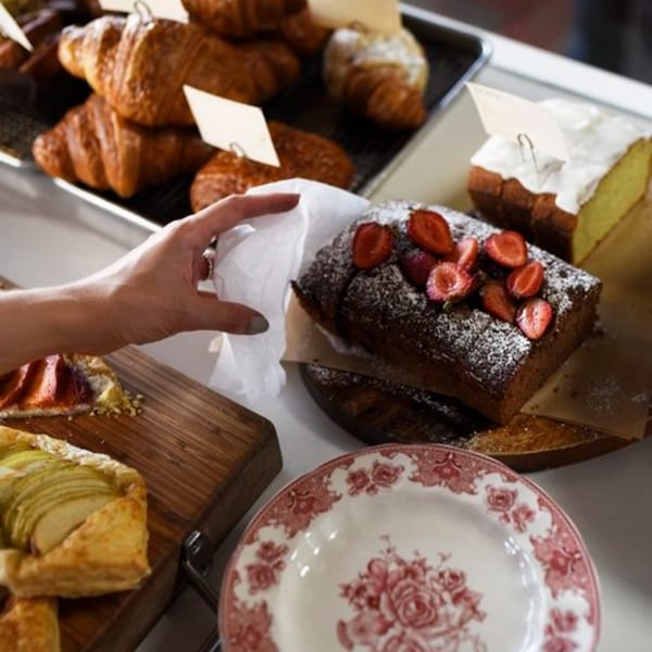 A hand reaching for a pastry on a counter filled with croissants, cakes, and tarts.